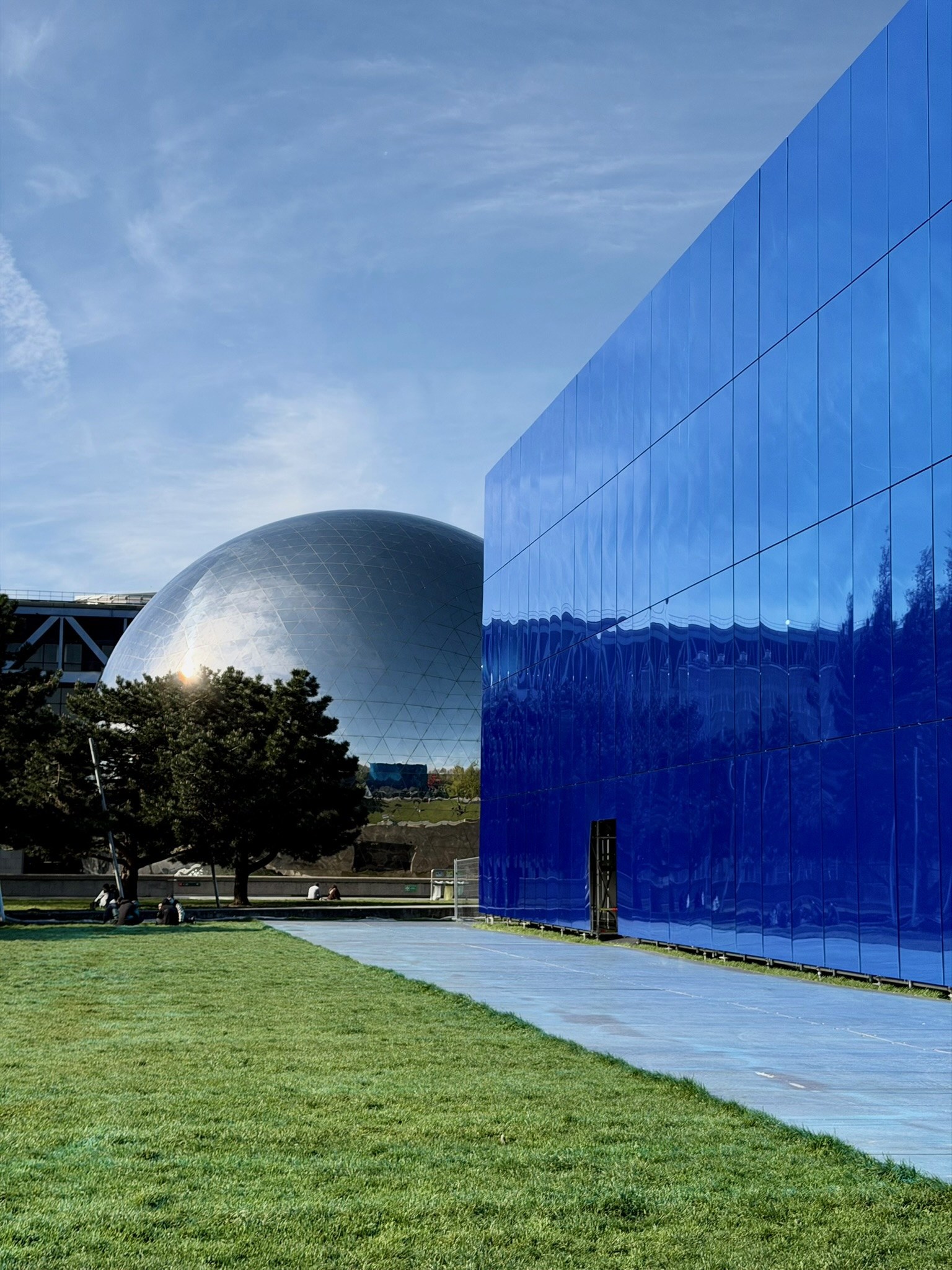 A mirrored steel sphere and a long wall of blue glass face each other above a lawn under a broken sky, photograph by Brandon Bisserier.